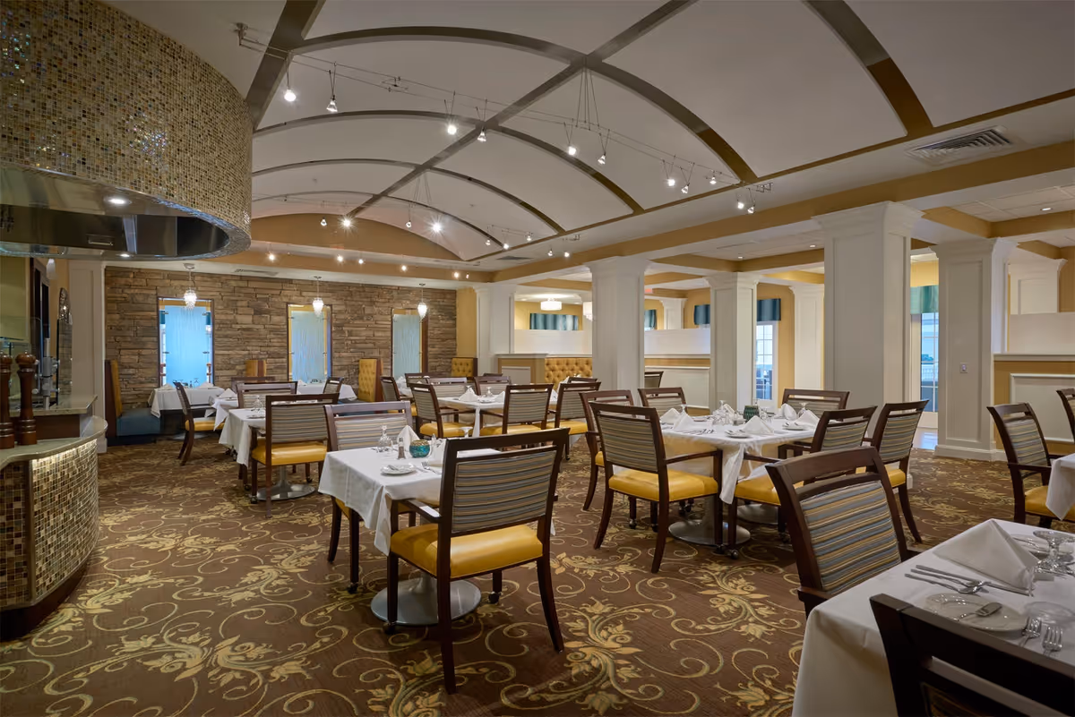 A spacious dining room with multiple tables covered in white tablecloths, set with glassware and napkins. The room features patterned carpet, cushioned chairs with wooden frames, stone accent walls, and a curved ceiling with modern lighting fixtures.