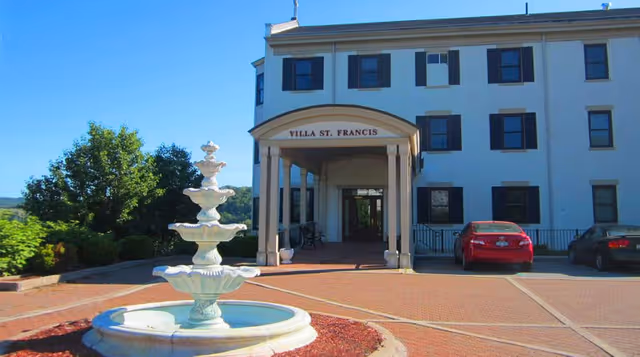 Front exterior view of Villa St. Francis building with a white multi-tiered fountain in the foreground, a brick-paved driveway, and some parked cars on the right side under a clear blue sky.