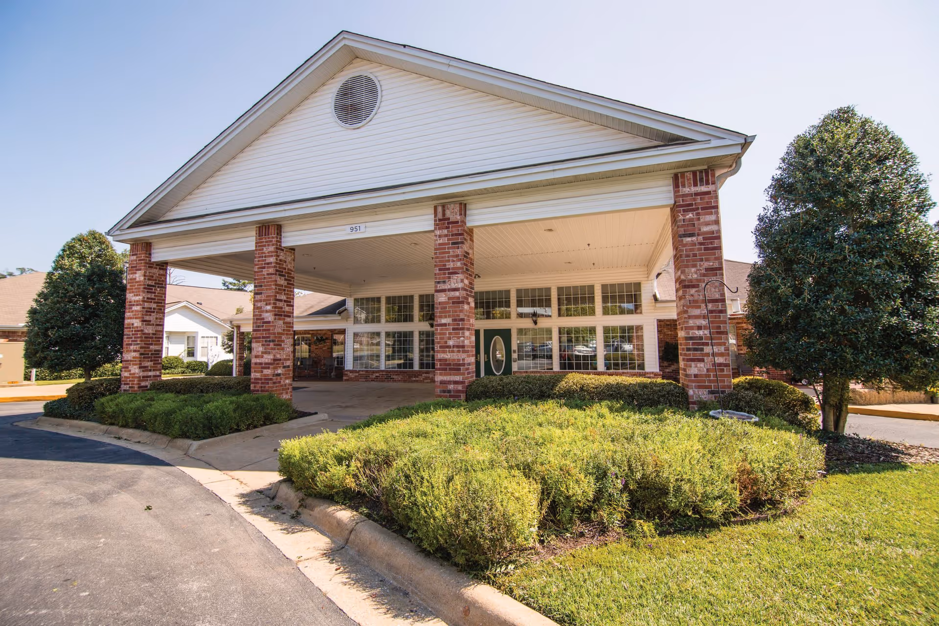 Front exterior view of a senior living facility with a covered entrance supported by brick columns, surrounded by neatly trimmed bushes and trees under a clear blue sky.