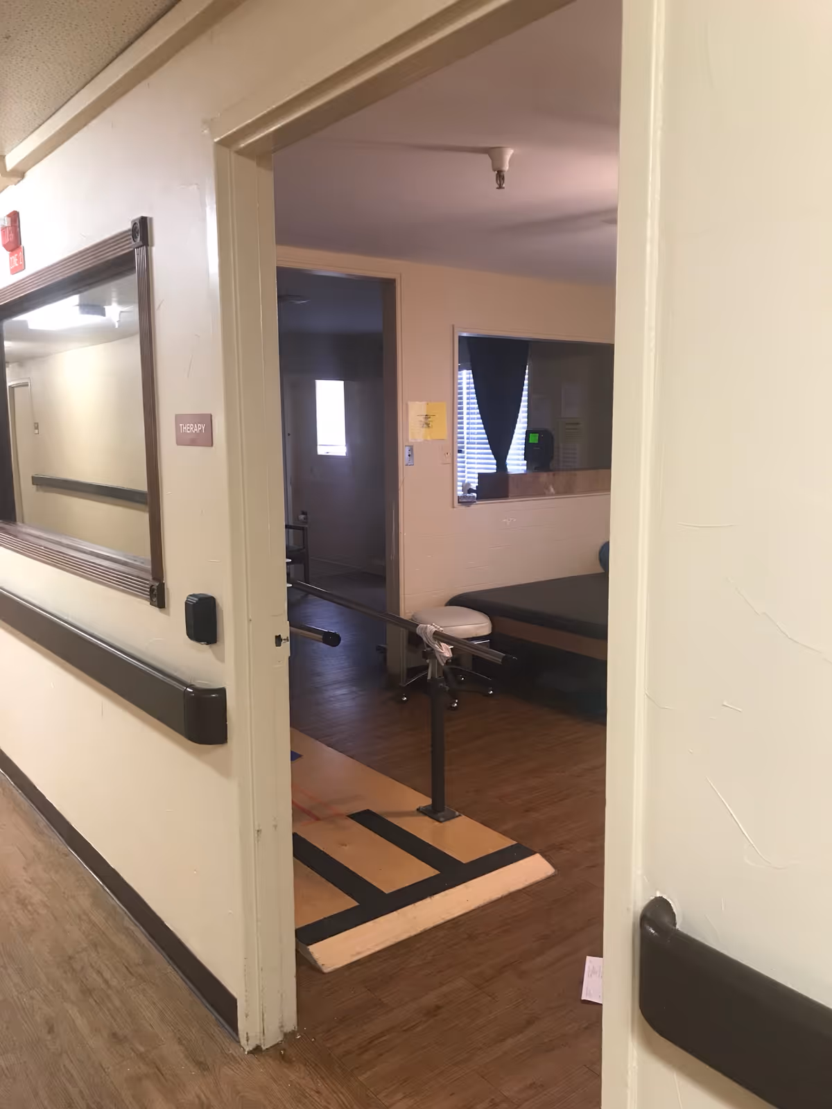 Hallway view into a rehabilitation therapy room with parallel walking bars, a treatment table, and wood-look flooring.