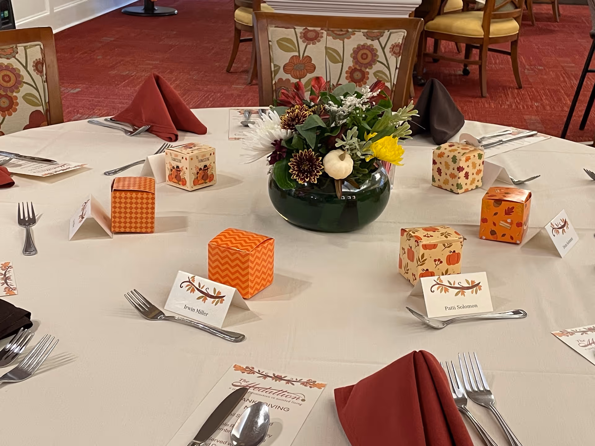 Round dining table set for a meal with a floral centerpiece, place cards, folded napkins and small decorative favor boxes.