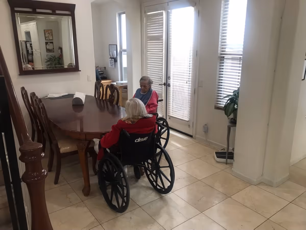 Two elderly women in wheelchairs sitting at a wooden dining table in a bright room with tiled floors, large windows with blinds, and a mirror on the wall.