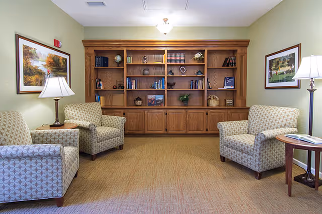 A cozy living room area with four patterned armchairs arranged around a wooden bookshelf filled with books and decorative items. Two table lamps with white shades are placed on side tables next to the chairs. The walls are light green and adorned with framed landscape paintings.