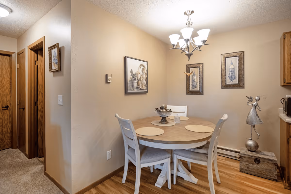 A cozy dining area with a round wooden table and four white chairs with cushions. The table has three round placemats and a decorative centerpiece. The walls are beige with three framed pictures hanging. A chandelier with five lights hangs above the table. To the right, there is a metallic sculpture of a girl holding a bird, standing on a wooden box. The floor is wooden, and there are three wooden doors visible on the left side of the image.