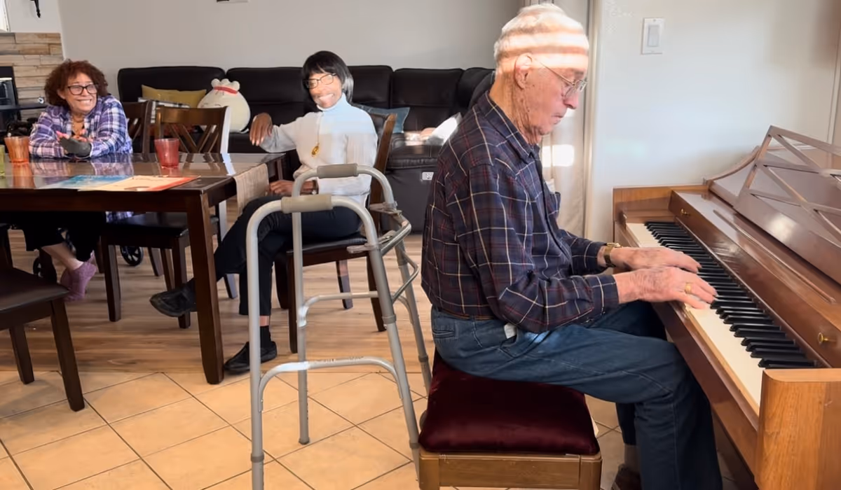 An elderly man playing a wooden piano in a living room area while two women sit at a dining table nearby, smiling and watching him. A walker is positioned near the man, and the room has tiled flooring and a dark sofa in the background.