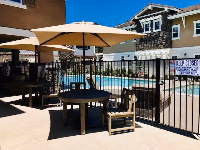 Outdoor seating area with round tables, chairs, and large beige umbrellas next to a fenced swimming pool at a residential community. In the background, there are multi-story buildings with white and brown exteriors under a clear blue sky.