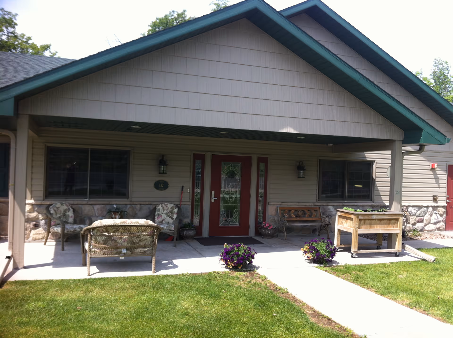 Front porch area of a building with a covered entrance, featuring a red door with decorative glass panels, two windows on either side, outdoor seating including cushioned chairs and a bench, a wooden planter box, and two flower pots with purple flowers on the concrete walkway leading to the entrance.