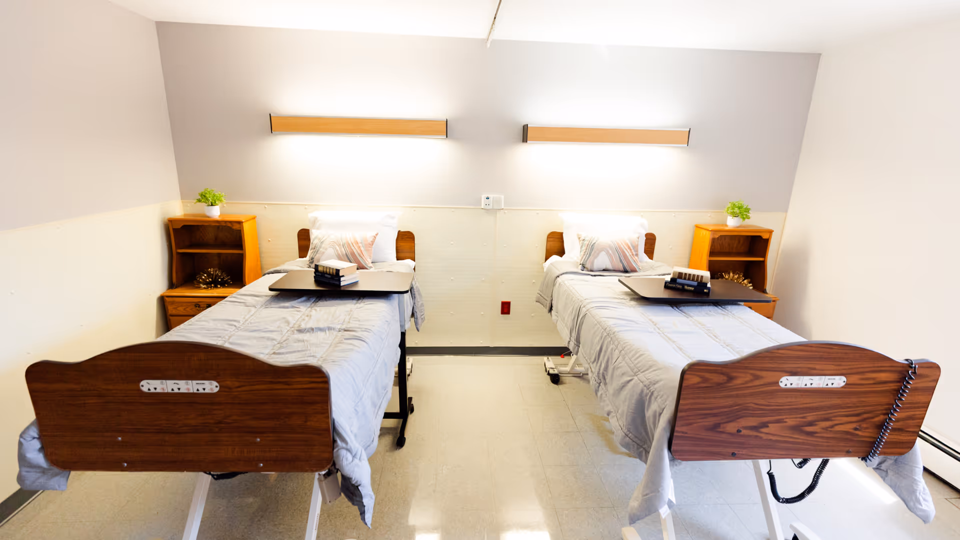A clean and simple room with two hospital-style beds, each with a gray blanket, a pillow, and a small overbed table holding books. Each bed has a wooden headboard and footboard, and there are two wooden nightstands with small green plants on top. The walls are light-colored with two horizontal wall lights above the beds.