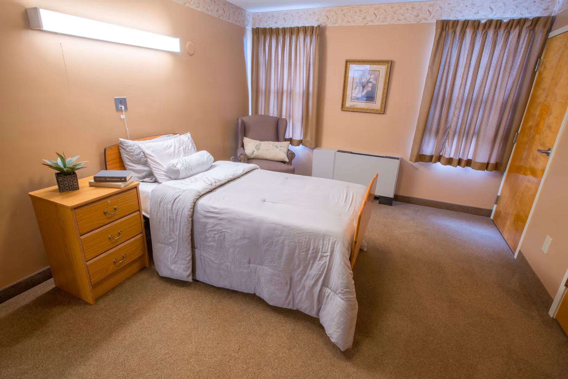 A neatly made single bed with white bedding in a cozy bedroom. Next to the bed is a wooden nightstand with a small potted plant and two books. There is a comfortable armchair with a decorative pillow near two windows covered with beige curtains. A framed picture hangs on the wall above a heating unit, and the room has beige walls and carpeted floor.
