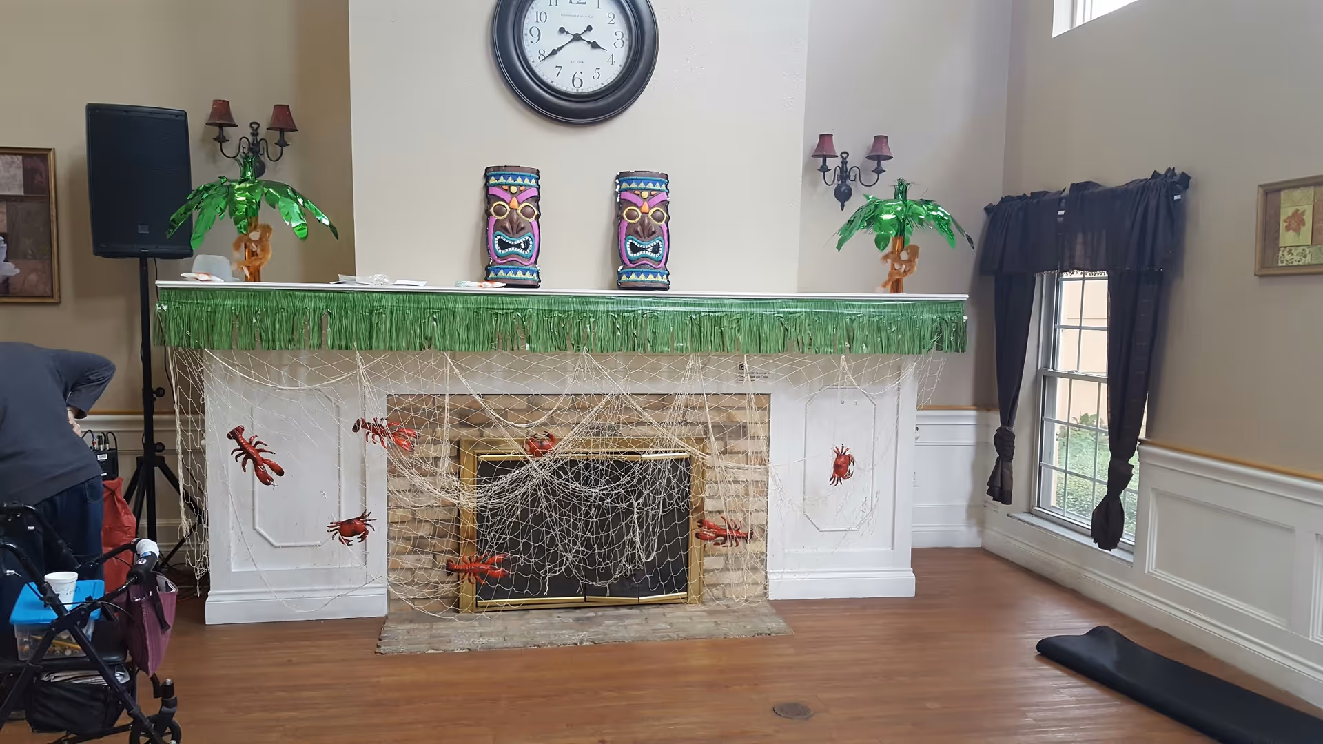 A decorated fireplace in a living room with a clock above it, two colorful tiki masks, and two small green palm tree decorations. The fireplace is adorned with a green fringe garland and a net with red lobster and crab decorations. There are wall sconces with red lampshades on either side of the fireplace and a window with dark curtains to the right.