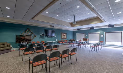 Spacious assisted-living activity room with rows of chairs facing a wall-mounted TV and projector under a recessed ceiling.