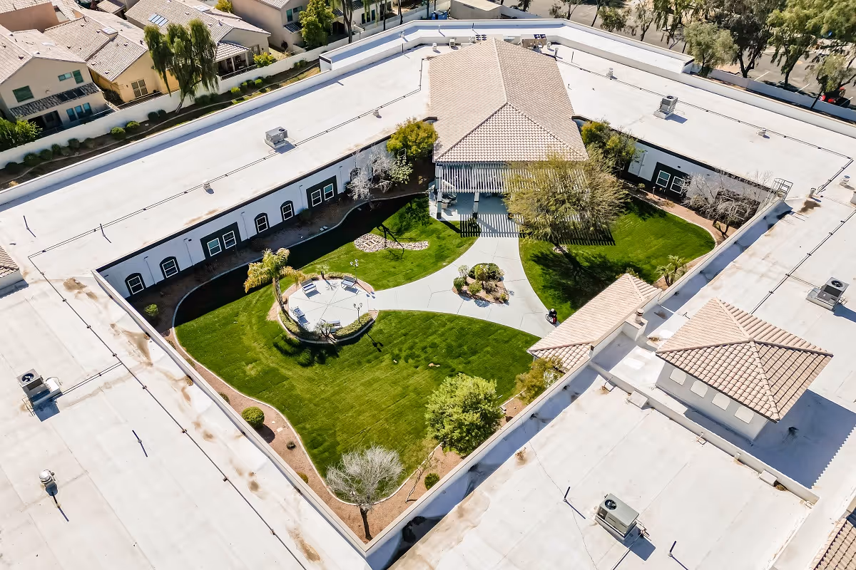 Aerial view of a senior living facility courtyard with a well-maintained green lawn, pathways, benches, palm trees, and surrounding buildings with tiled roofs.