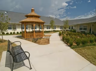 Outdoor courtyard area with a wooden gazebo in the center, surrounded by a paved walkway, landscaped greenery, small trees, and a black metal bench on the left side. The courtyard is enclosed by a single-story building under a partly cloudy sky.