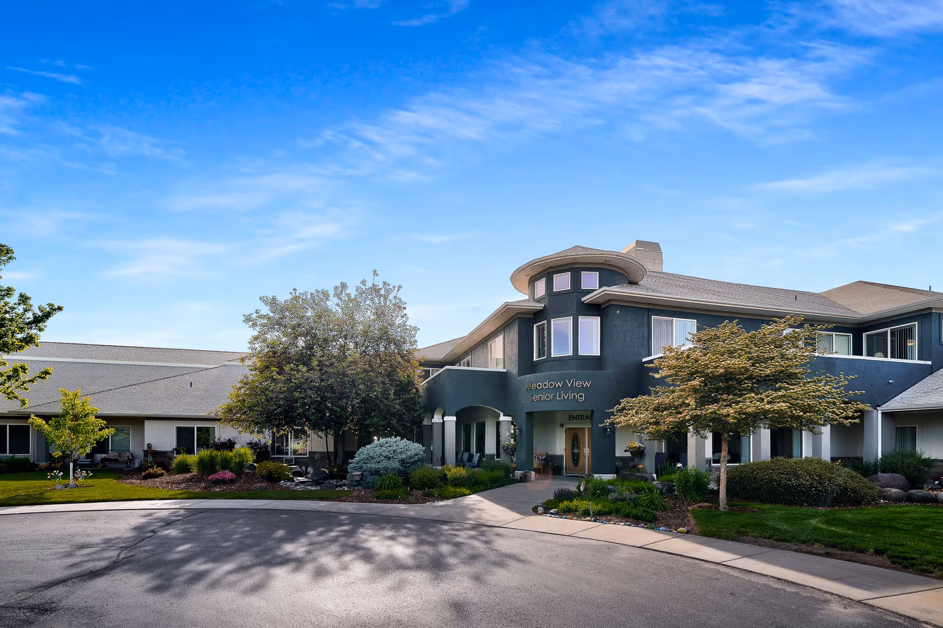 Exterior view of Meadow View Senior Living facility showing a two-story building with a rounded turret-like entrance, surrounded by well-maintained landscaping including trees, shrubs, and a lawn under a clear blue sky.