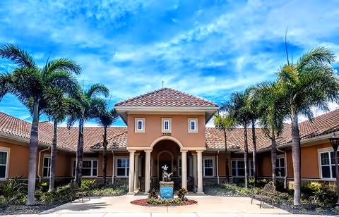 Exterior view of Gulf Coast Memory Care facility featuring a central entrance with a tiled roof, flanked by palm trees and benches under a partly cloudy blue sky.
