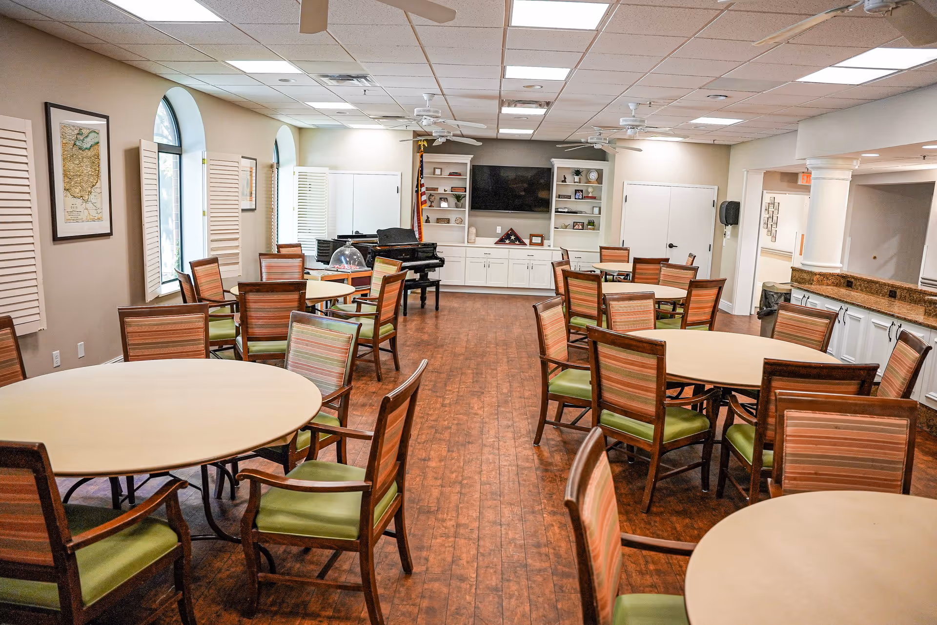A spacious senior living facility common area with multiple round tables surrounded by wooden chairs with green cushions. The room has wood flooring, beige walls, and several arched windows with white shutters. At the far end, there is a black piano, a large flat-screen TV mounted on the wall, and built-in white cabinets with shelves displaying decorative items. Ceiling fans and recessed lighting are visible on the ceiling.