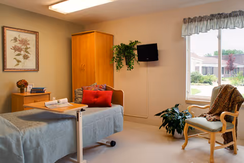 A cozy room with a single bed covered in a gray blanket and a red pillow. Next to the bed is a wooden nightstand with a small plant and some books. A wooden wardrobe stands against the wall, and a flat-screen TV is mounted above it. A window with a valance lets in natural light, showing a view of a garden outside. A wooden chair with a cushion and a blanket is placed near the window, next to a green potted plant.