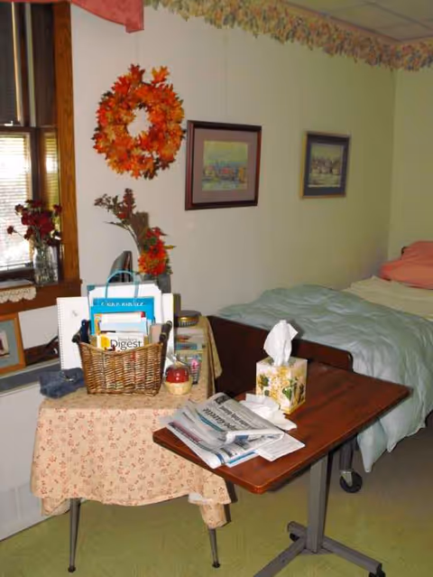 A cozy nursing home bedroom with a bed, table, magazines, tissue box, and autumn wreath.