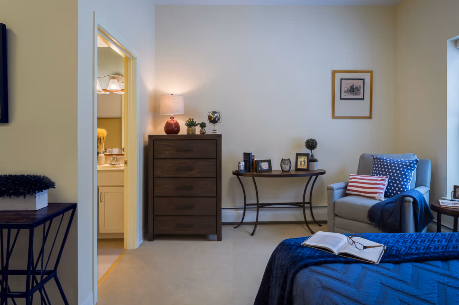 A cozy bedroom in a senior living facility with a bed covered in a blue quilt and an open book with glasses on it. There is a gray armchair with patriotic-themed pillows and a blue throw blanket next to a small round side table. A wooden dresser with a red lamp, small plants, and a globe sits against the wall. A narrow console table holds framed photos and decorative items. The bathroom door is open, showing a sink and mirror with lights above it.