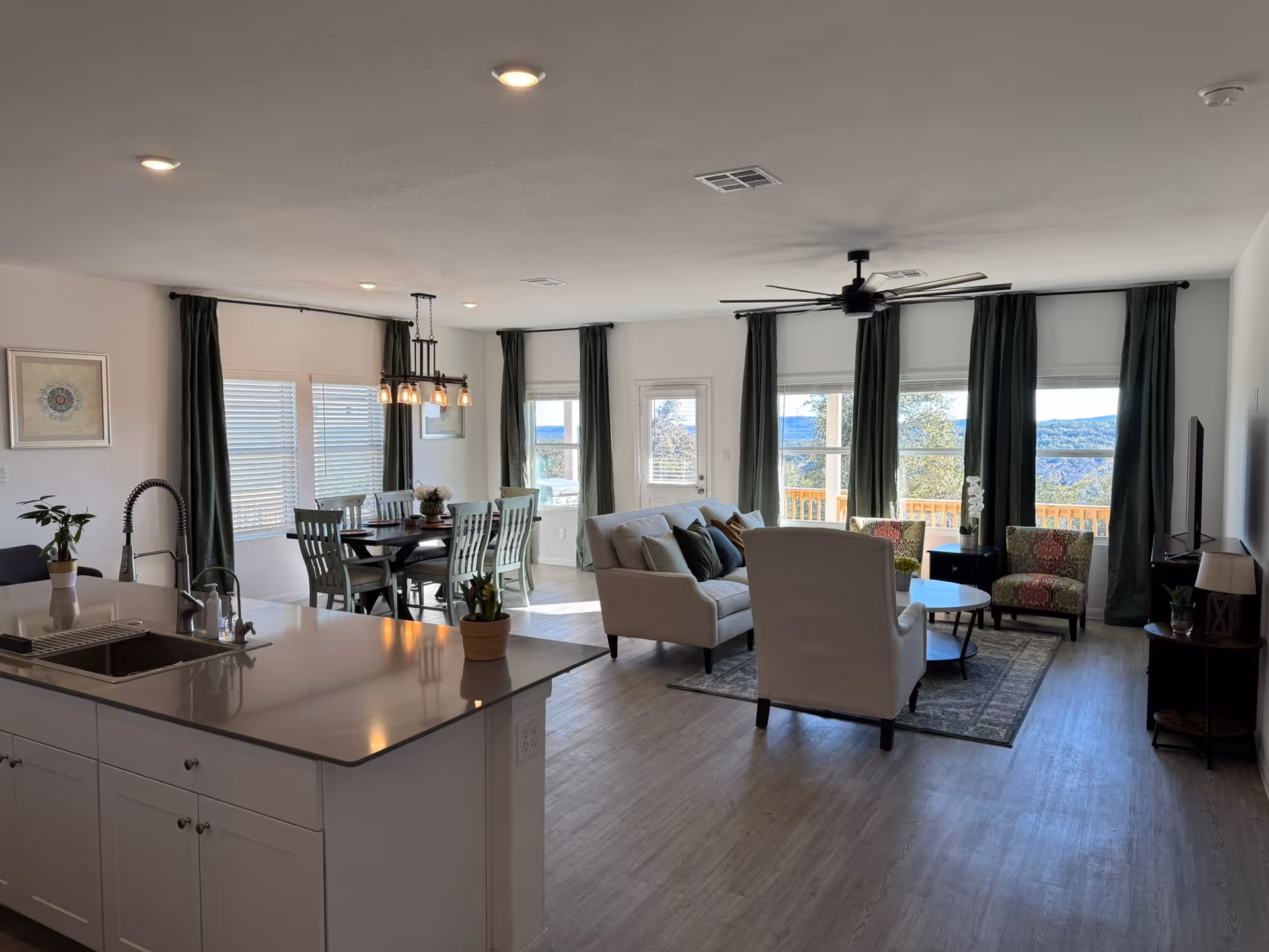 A bright and spacious open-concept living area featuring a modern kitchen island with a sink and faucet in the foreground, a dining table with six chairs and a hanging light fixture in the midground, and a cozy living room with a sofa, armchair, two patterned chairs, a coffee table, and a TV on a stand near large windows with dark green curtains overlooking a scenic outdoor view.