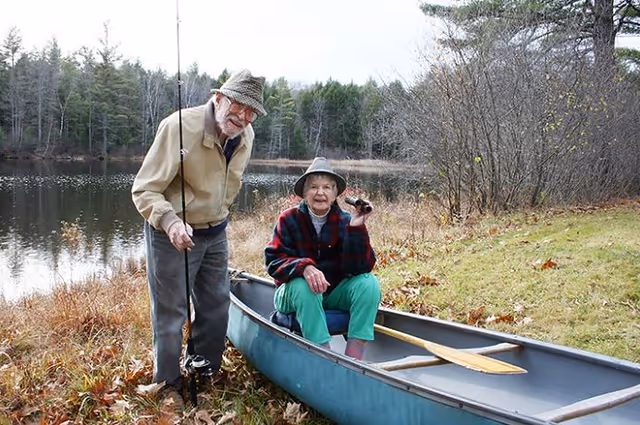 An elderly man and woman outdoors near a lake. The man is standing on the grassy shore holding a fishing rod, wearing a beige jacket, gray pants, and a hat. The woman is sitting in a blue canoe on the grass, wearing a plaid jacket, green pants, and a hat, holding binoculars. Trees and water are visible in the background.