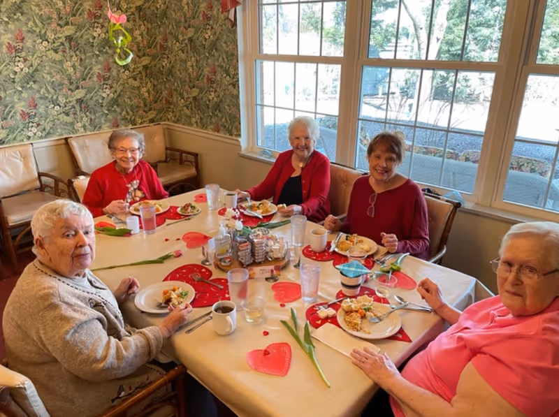 Six senior women sitting around a decorated dining table enjoying a meal in a bright dining room with large windows.