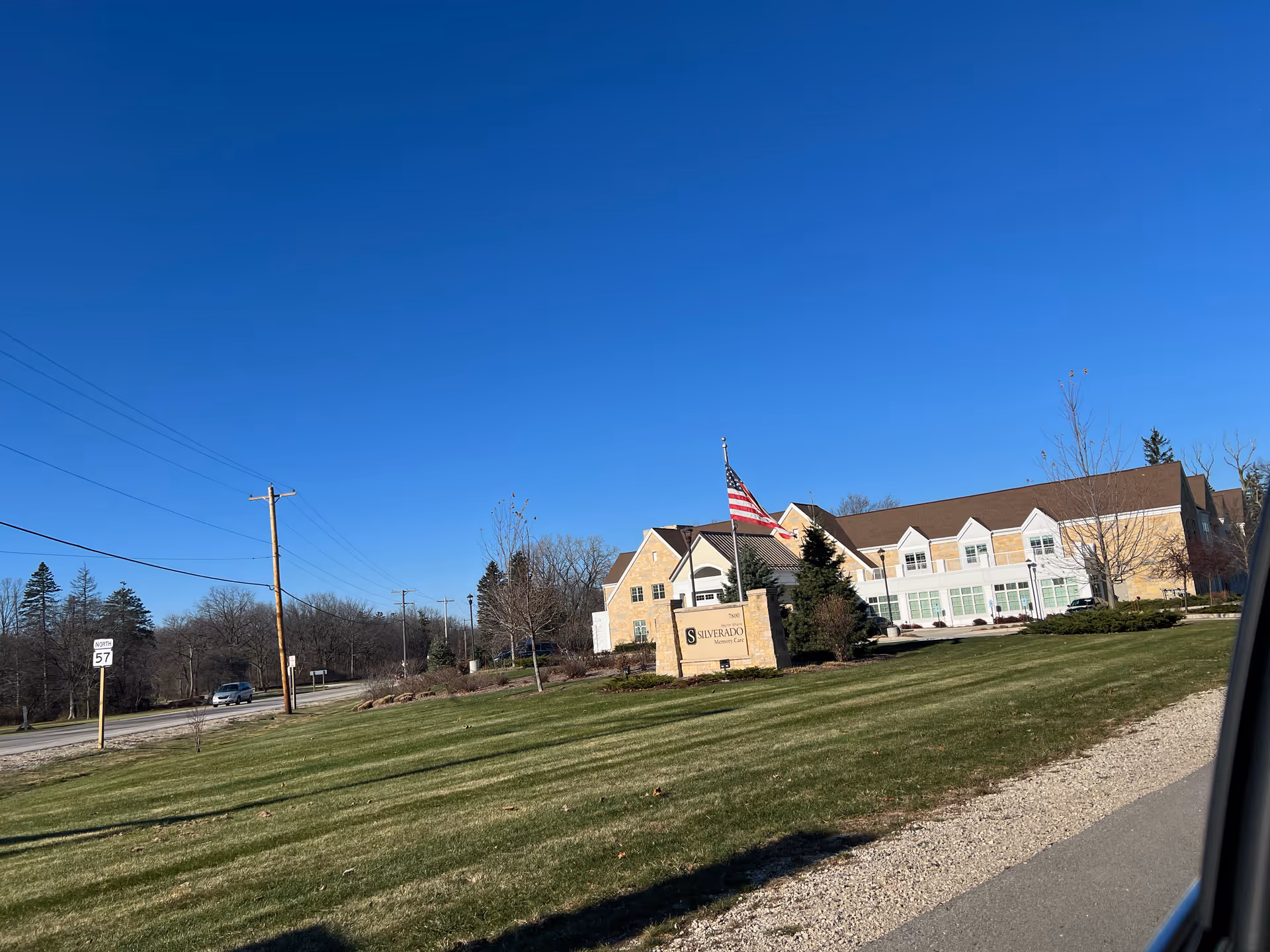 Exterior view of Silverado North Shore Memory Care Community building with a large green lawn, an American flag on a flagpole, and a clear blue sky.