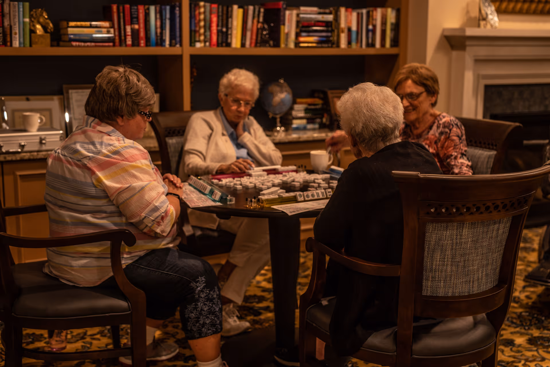 Four elderly women sitting around a table playing a game of Mahjong in a cozy room with bookshelves and a fireplace in the background.