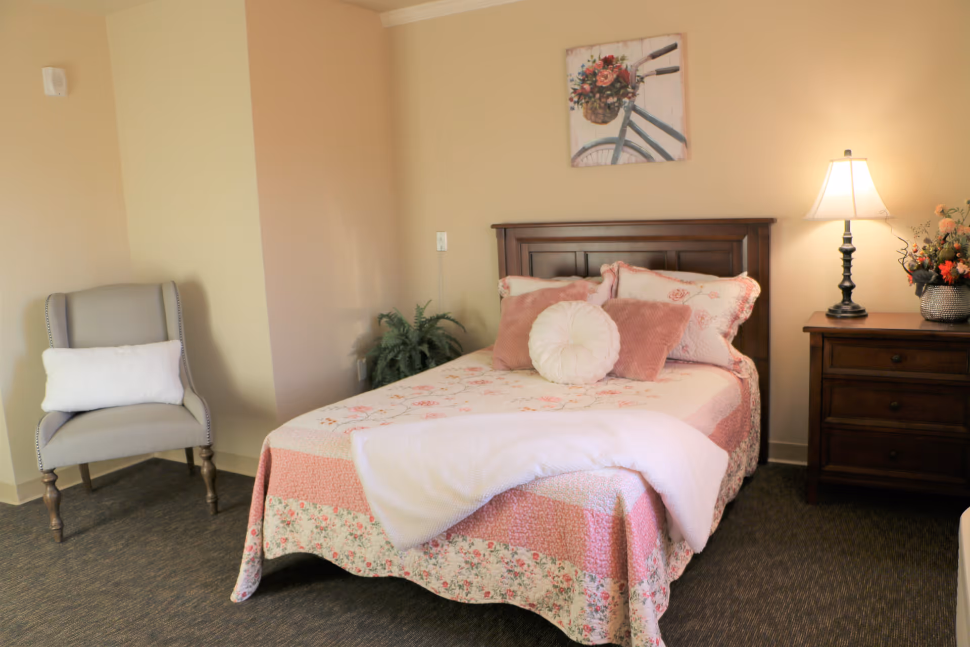 A cozy bedroom with a wooden bed frame and a floral pink and white quilt. The bed is decorated with multiple pillows including a round white one and two pink ones. To the right of the bed is a wooden nightstand with a lit table lamp and a vase of flowers. On the left side of the room, there is a gray upholstered armchair with a white pillow. A painting of a bicycle with a basket of flowers hangs on the wall above the bed.
