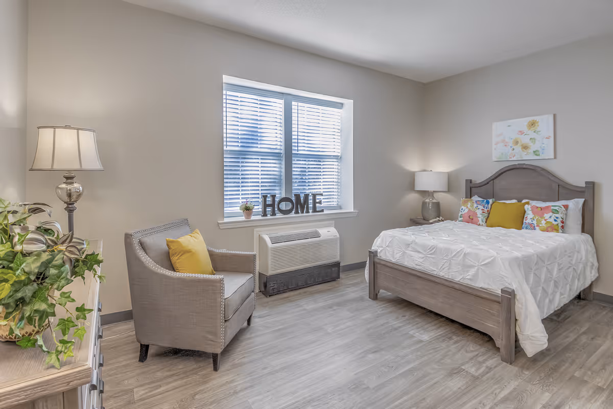 A bright and cozy bedroom with a wooden bed featuring white bedding and colorful floral pillows. There is a gray armchair with a yellow cushion next to a window with blinds. The window sill has a small potted plant and decorative letters spelling 'HOME'. A wooden dresser with a lamp and a green leafy plant is on the left side. The room has light-colored walls and wood flooring.