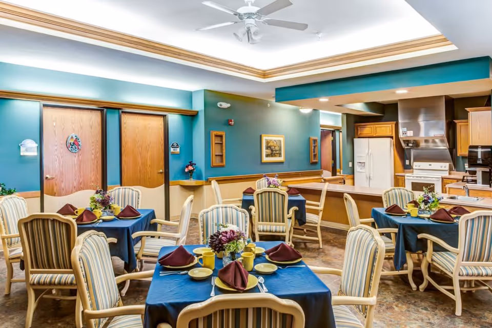 Dining room with set tables, striped armchairs, floral centerpieces and an open kitchen area in the background.