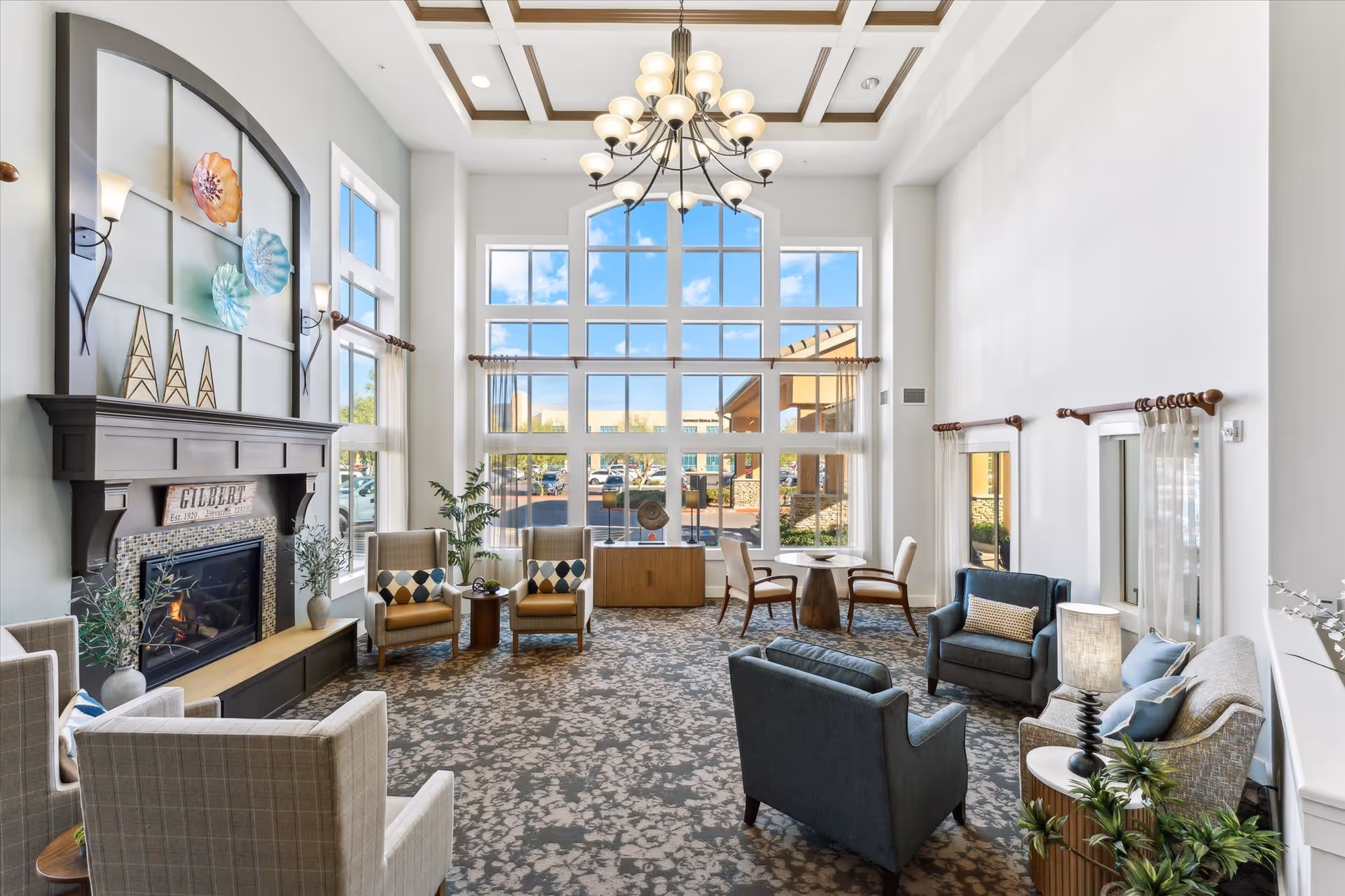 A spacious and well-lit living room area with large floor-to-ceiling windows showing a view of the outside parking lot. The room features a fireplace with decorative art above it, multiple comfortable armchairs and sofas arranged around small tables, a chandelier hanging from a coffered ceiling, and plants adding greenery to the space.