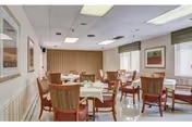 A dining room with several tables covered with white tablecloths and surrounded by wooden chairs with cushioned seats and backs. The room has large windows with blinds, framed artwork on the walls, and a ceiling with fluorescent lighting.