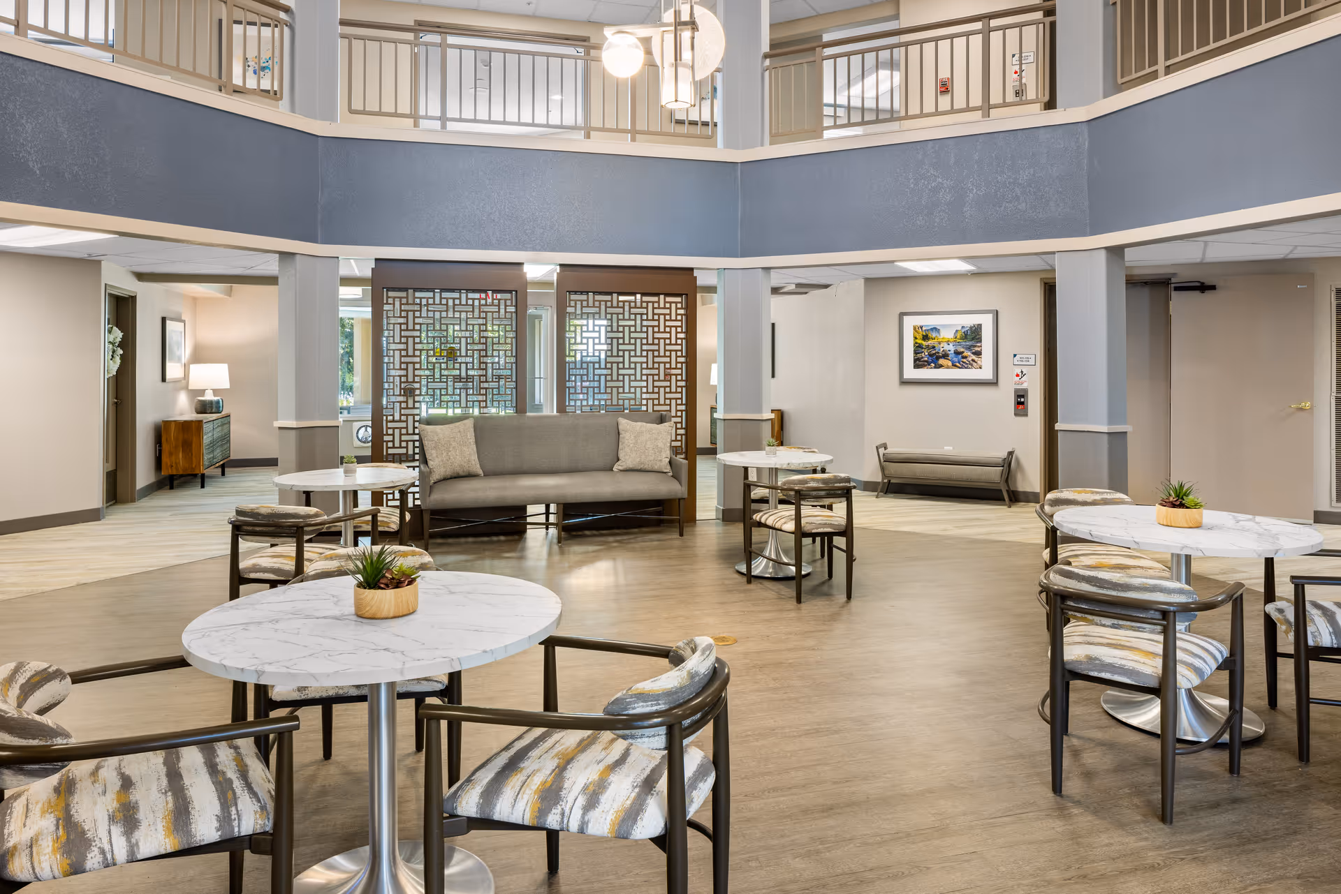 A spacious senior living community common area with round marble-top tables and cushioned chairs arranged around the room. A gray sofa with two pillows is positioned against a decorative wooden screen. The room has light wood flooring, neutral-colored walls, and a second-floor balcony railing visible above. There are potted plants on the tables and framed artwork on the walls.