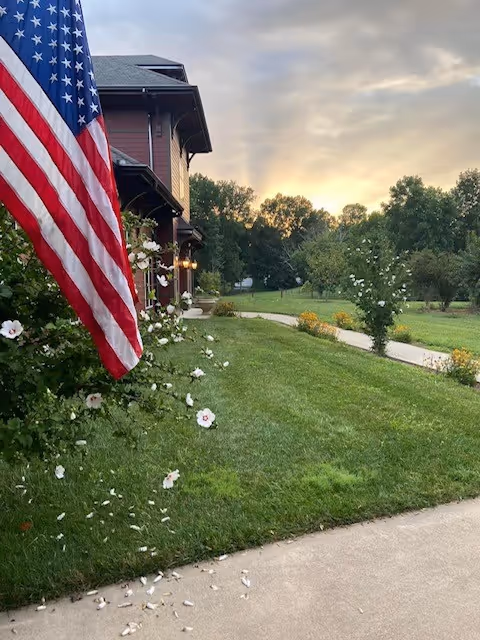 View of a green lawn with flowering bushes and a concrete pathway leading towards a building with a porch. An American flag is prominently displayed on the left side near the building. Trees and a cloudy sky are visible in the background.