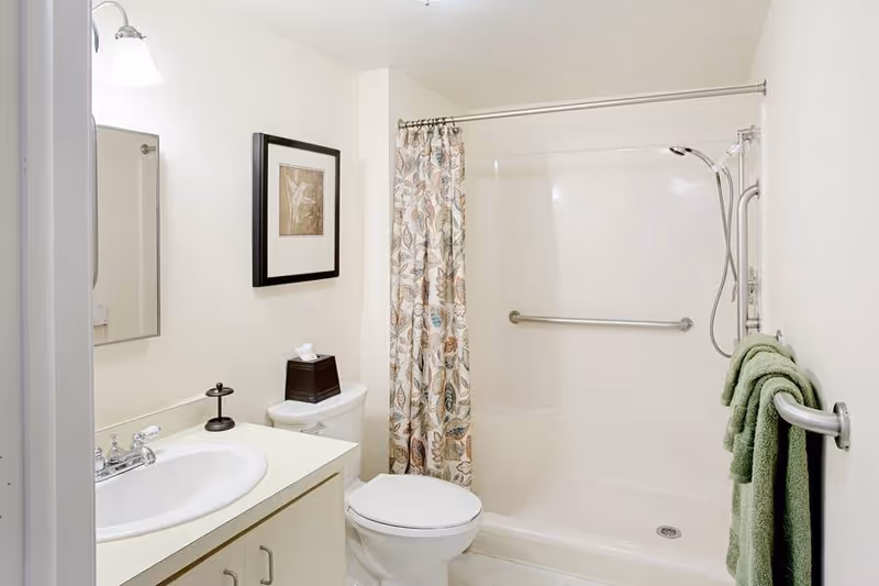 A clean and bright bathroom featuring a white sink with a silver faucet, a toilet, a shower with a floral-patterned curtain, and green towels hanging on a silver towel rack. There is a framed picture on the wall above the toilet and a tissue box on the toilet tank.