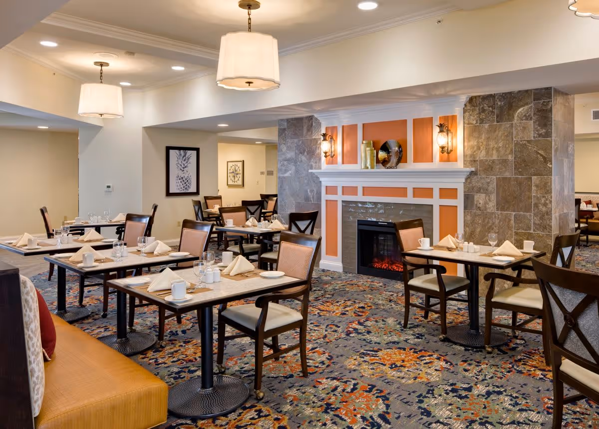 A dining room in a senior living facility with multiple tables set with napkins, plates, cups, and glasses. The room features a decorative fireplace with orange and white paneling, stone accents, and wall sconces. There are cushioned chairs around the tables and a patterned carpet covering the floor. Pendant lights hang from the ceiling, and framed artwork is visible on the walls.