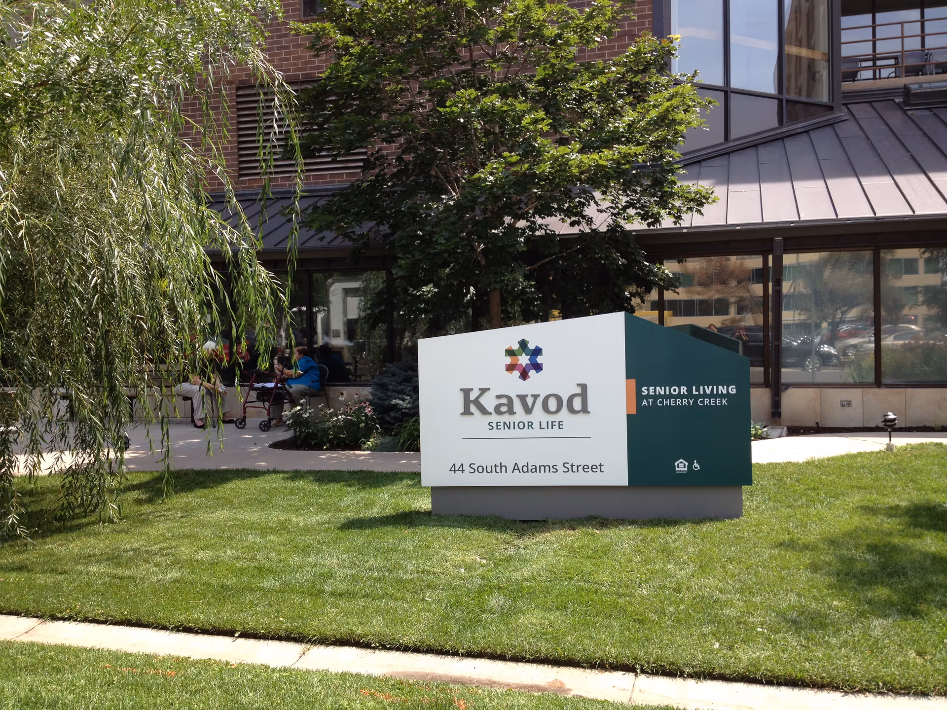 Outdoor view of the Kavod Senior Life facility sign on a grassy area with trees and a building with large windows in the background. Two people are sitting on a bench near the building.