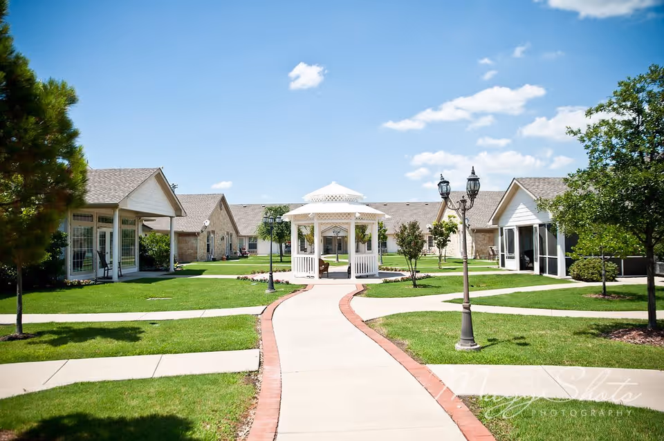 A bright outdoor courtyard at Bethesda Gardens Frisco Assisted Living featuring a paved walkway with red brick edging leading to a white gazebo in the center. The area is surrounded by well-maintained green lawns, trees, lamp posts, and single-story buildings with beige walls and pitched roofs under a blue sky with scattered clouds.
