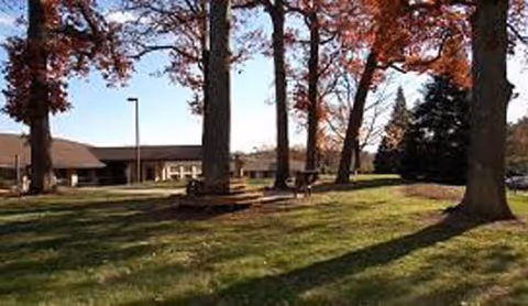 Outdoor area with large trees casting shadows on a grassy lawn in front of a single-story building under a clear sky.