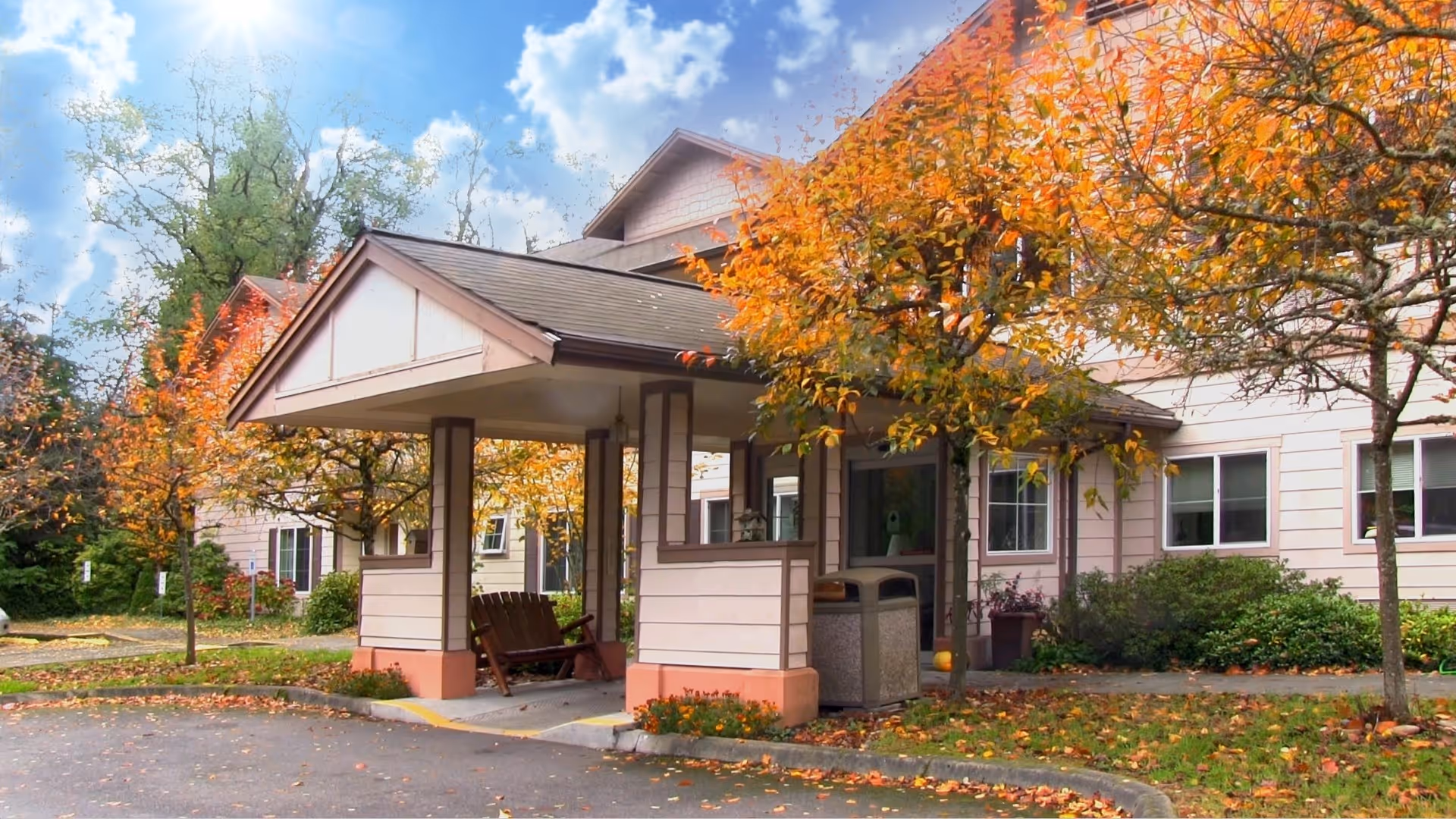 Exterior view of a senior living facility entrance with a covered drop-off area, surrounded by trees with autumn foliage and a clear blue sky with some clouds.