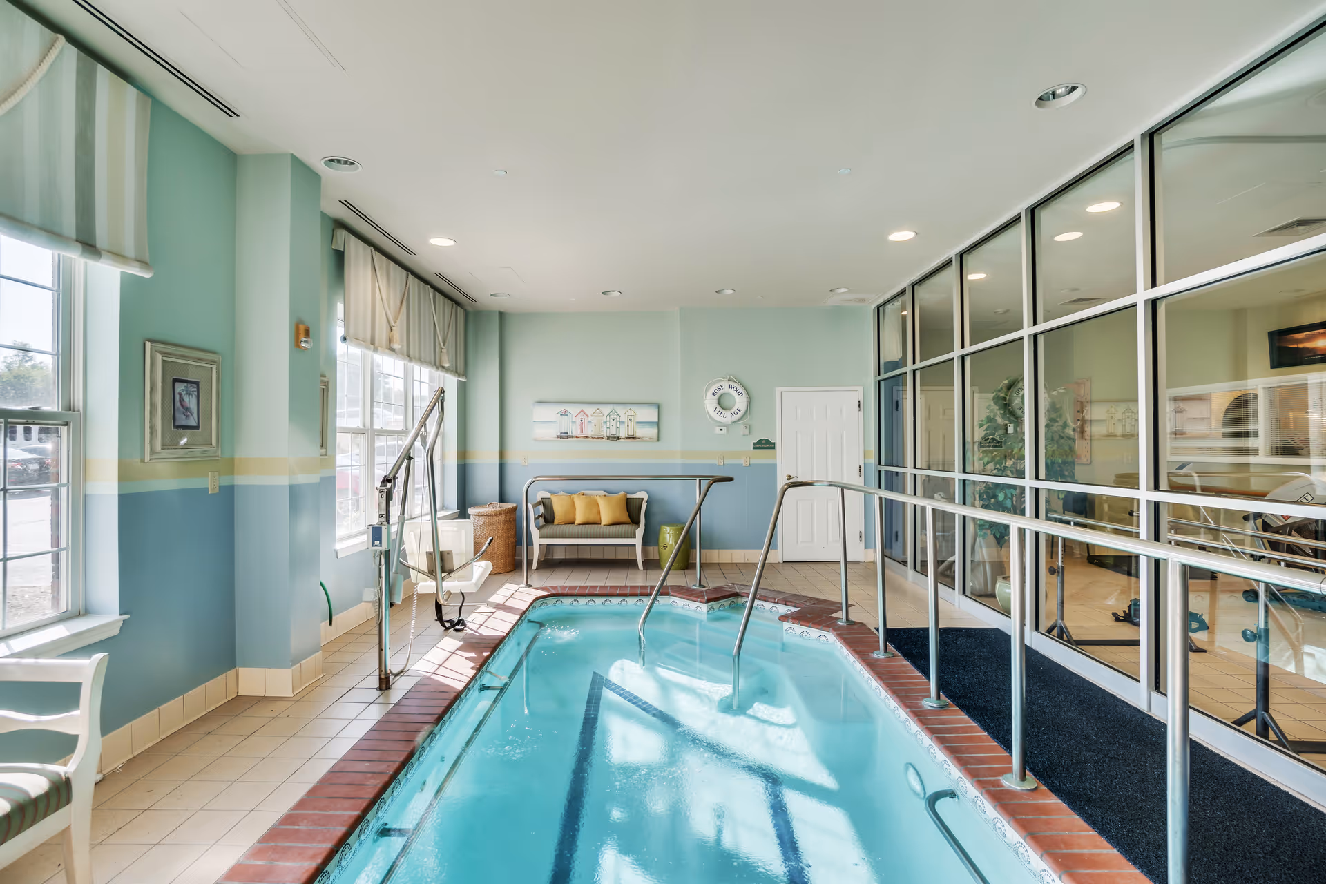 Indoor therapeutic pool with handrails, a pool lift, seating and large windows in a bright aquamarine room.