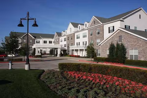 Sunlit exterior view of Prestonwood Court senior living facility with manicured landscaping and driveway.