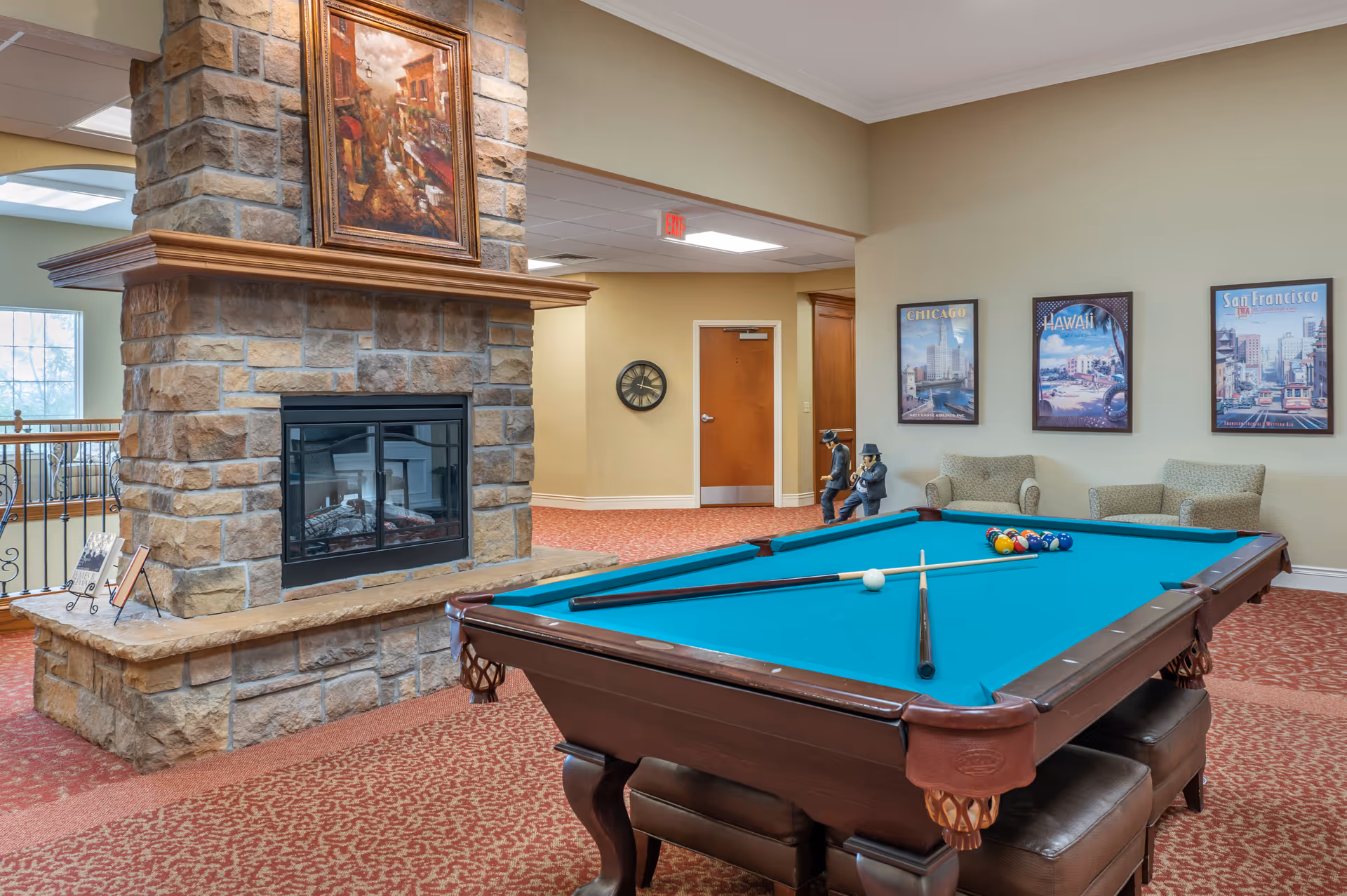 A cozy indoor recreational room featuring a pool table with cues and balls set up, a stone fireplace with a painting above it, two armchairs, and three framed travel posters on the wall labeled Chicago, Hawaii, and San Francisco. The room has warm lighting and patterned carpet.