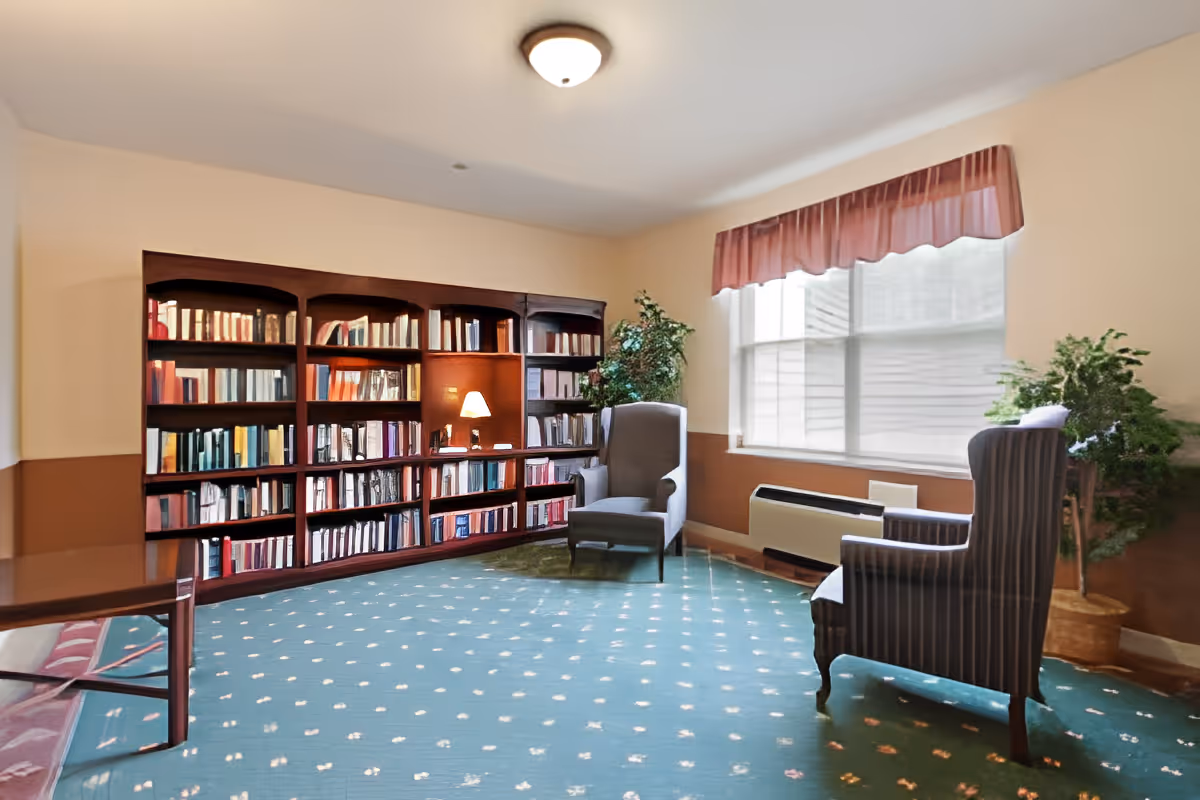 Sunlit reading room with wall-length bookshelves, two upholstered armchairs, and a large window.