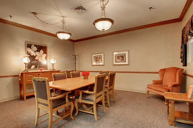 A dining room with a wooden table surrounded by six chairs with striped upholstery. The room features beige walls with a wooden chair rail and crown molding. There are two framed pictures on the wall, a sideboard with two lamps and a floral painting above it, a coat rack, and an upholstered armchair in the corner next to a small wooden side table. Two ceiling light fixtures hang above the table.