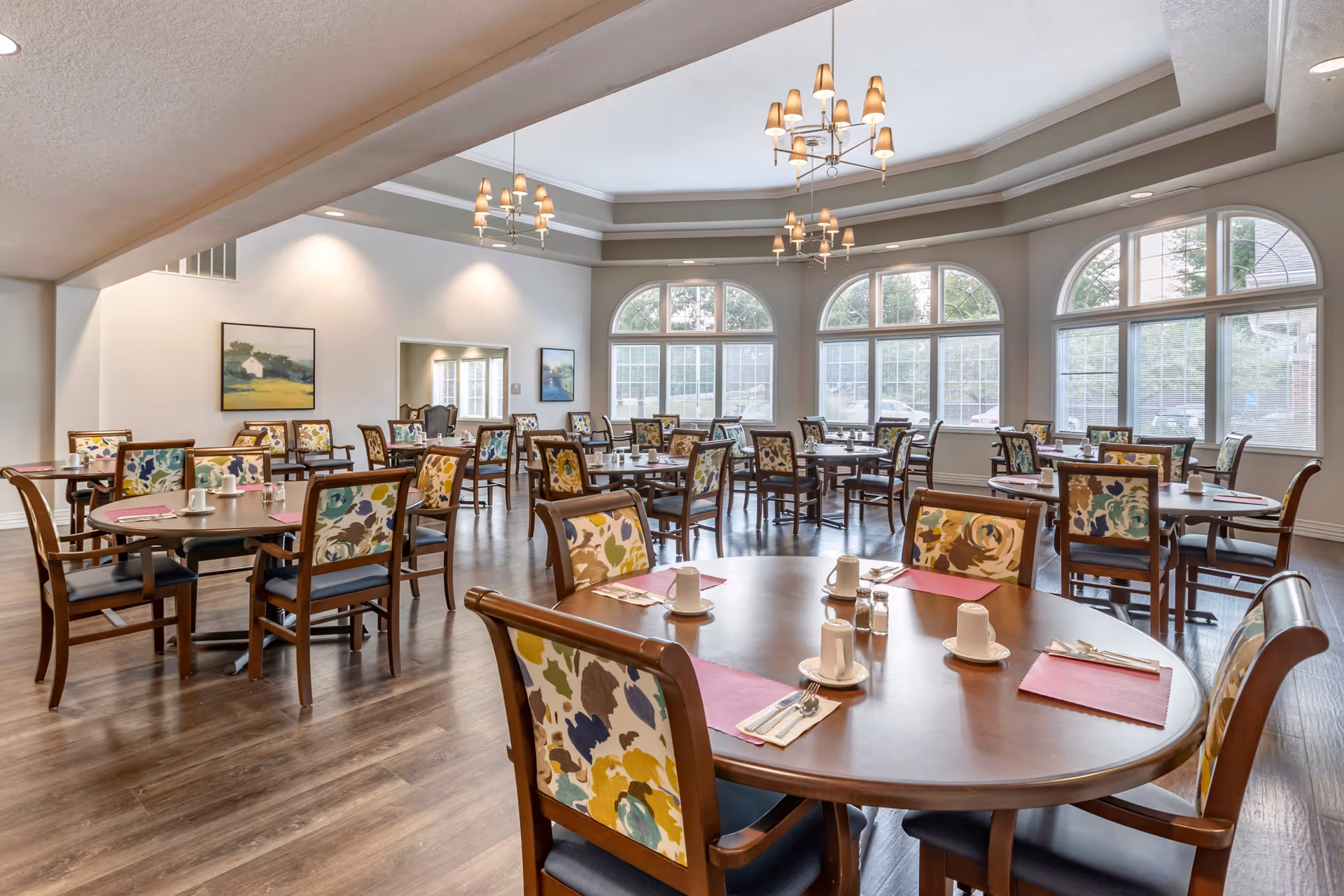 A spacious dining room in a senior living facility with multiple round wooden tables and chairs featuring floral upholstery. Each table is set with white cups, saucers, silverware, and pink placemats. Large arched windows let in natural light, and elegant chandeliers hang from the ceiling. The room has light-colored walls, wooden flooring, and framed artwork on the walls.
