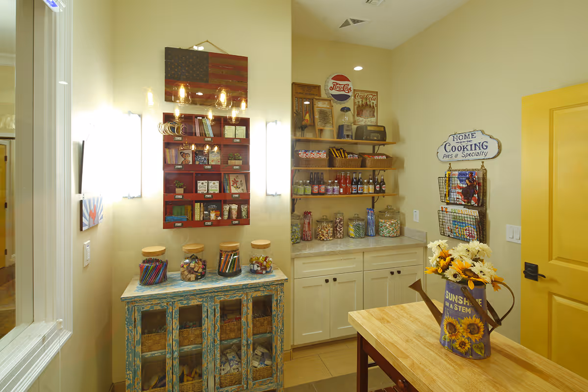 A cozy room with a rustic blue cabinet holding jars filled with colorful craft supplies, a red wall-mounted organizer with various small items, and shelves stocked with snacks and beverages. A yellow door is visible on the right, and a wooden table with a watering can vase holding sunflowers is in the foreground. The walls are light yellow, and there is a sign that reads 'Home Cooking pies & Specialty' along with a magazine rack.