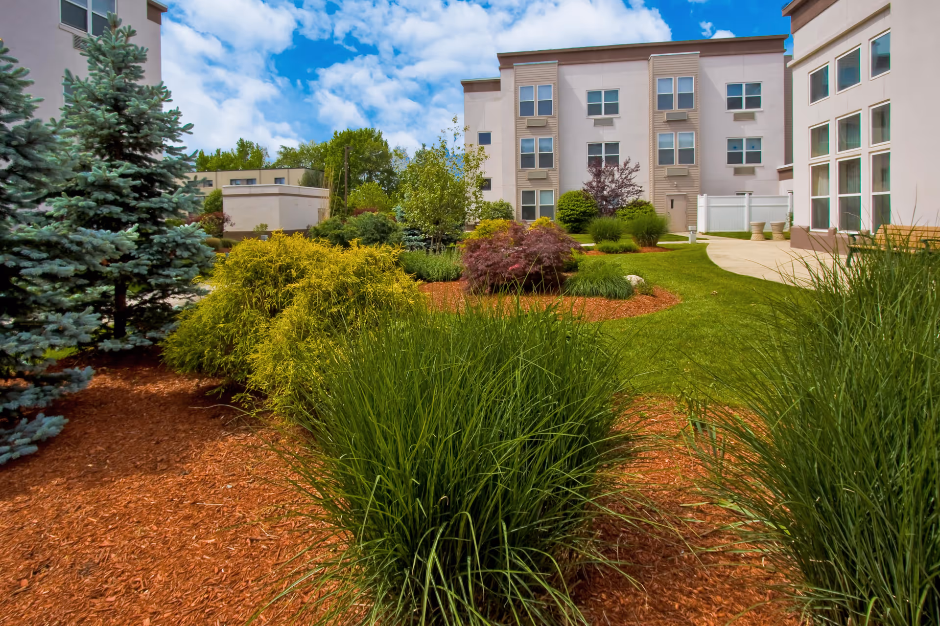 A landscaped outdoor garden area with various green shrubs, bushes, and trees surrounded by red mulch. In the background, there is a multi-story building with multiple windows and a clear blue sky with some clouds.