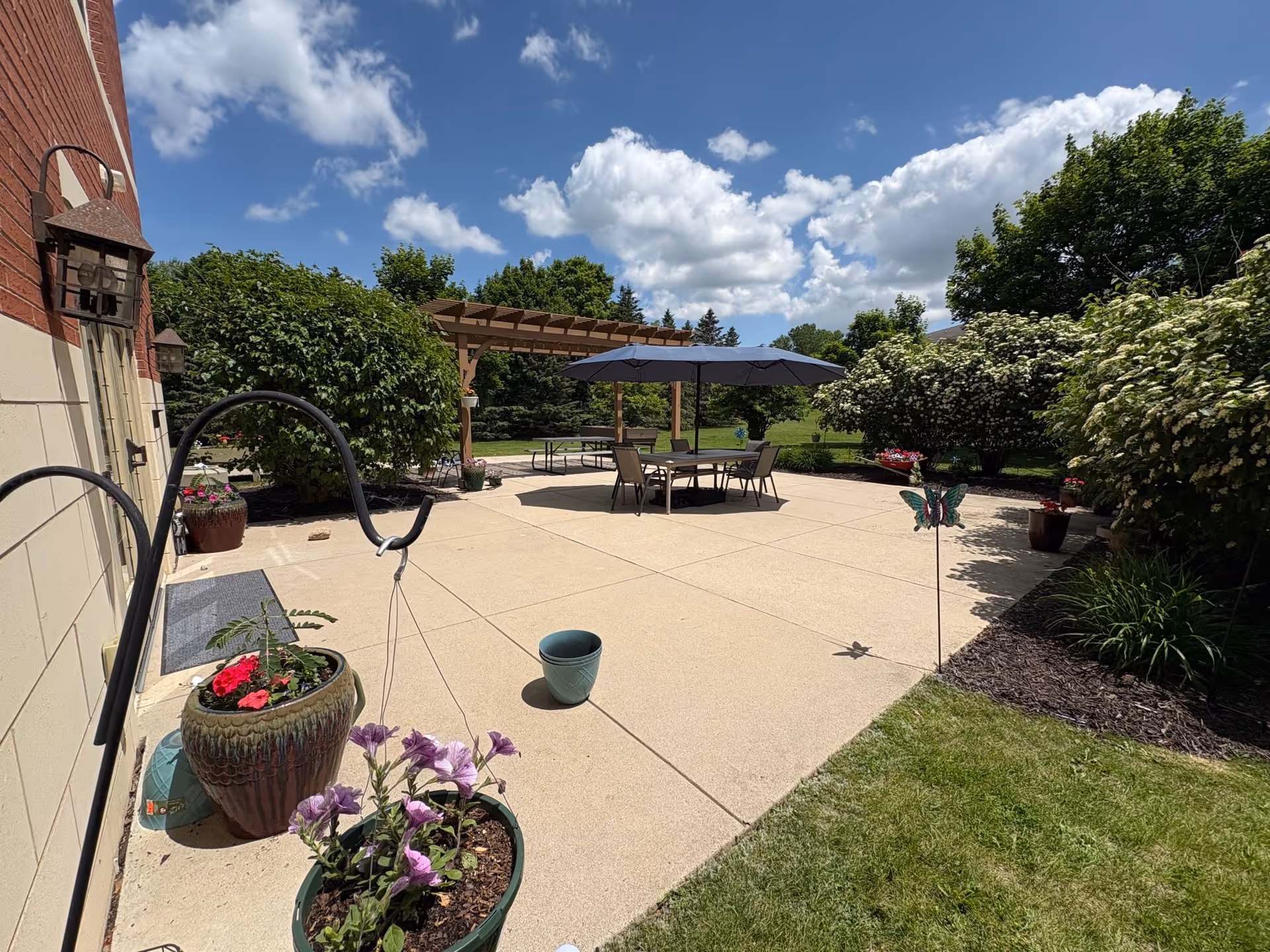 Outdoor patio area with a large concrete surface, several potted plants, a table with chairs under a large umbrella, and a wooden pergola in the background. The area is surrounded by green bushes and trees under a partly cloudy blue sky.